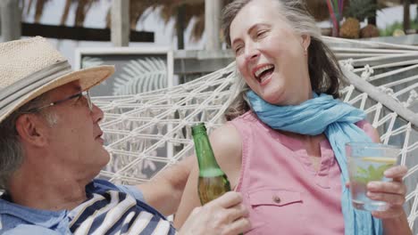 Glückliches-älteres-Kaukasisches-Paar,-Das-In-Zeitlupe-Auf-Der-Hängematte-Vor-Der-Strandbar-Trinkt-Und-Redet