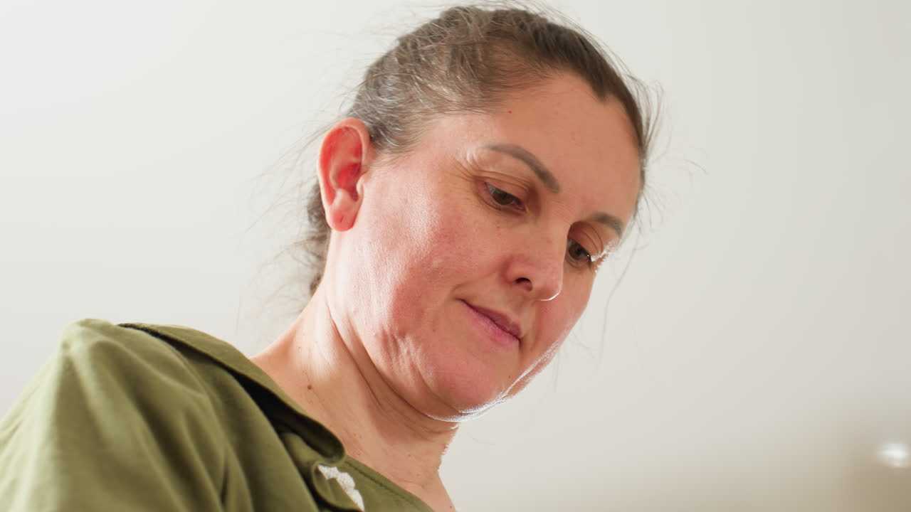 Middle age woman in green blouse looks down with focused expression while gently scratching face, appearing thoughtful and observant in bright kitchen setting