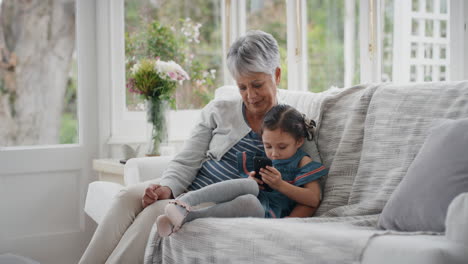 Entzückendes-Kleines-Mädchen,-Das-Großmutter-Zeigt,-Wie-Man-Ein-Smartphone-Benutzt,-Und-Oma-Moderne-Technologie-Beibringt,-Intelligentes-Kind,-Das-Oma-Mit-Dem-Mobiltelefon-Zu-Hause-Hilft,-4K