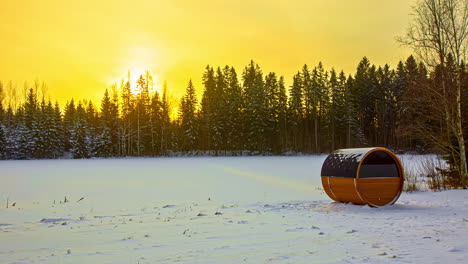 Barrel-Sauna-On-Snowy-Field-With-Coniferous-Forest-In-Background-And-Bright-Yellow-Sunset-Sky