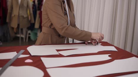 Professional-female-tailor-is-working-at-table-with-red-cloth-in-atelier.-Skilled-seamstress-is-drawing-sketch-lines-with-a-soap-or-a-chalk-on-red-fabric-using-patterns.-Close-up