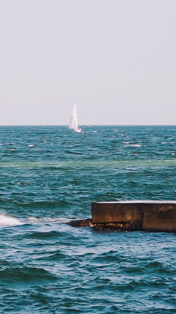 Motion of sea water. Waves break at stone pier and make white foam. Beautiful blue sea and white sail floating on alone. Slow motion. Vertical video