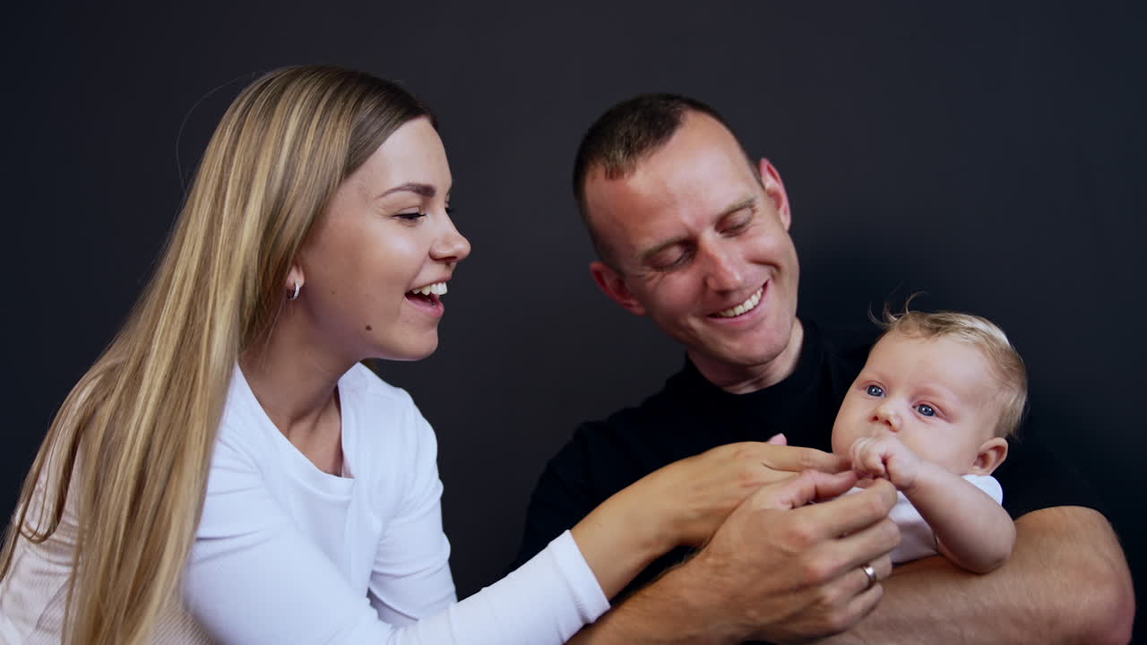 Lovely blonde infant sitting in dad's hands. Mother plays with her adorable son. Black backdrop.