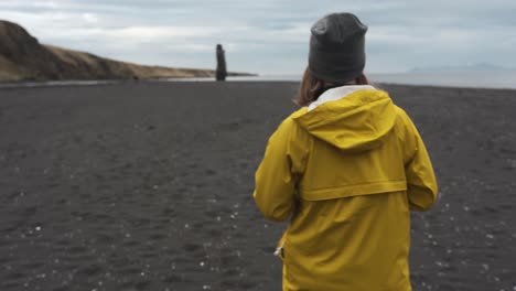 mujer con un impermeable amarillo caminando en una playa de arena negra en hvitserkur, vatnsnesvegur ubicado en islandia