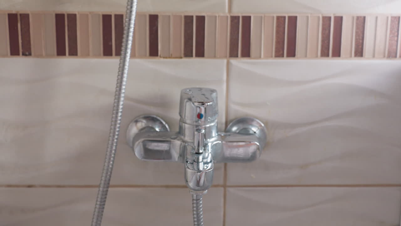 Bathroom scene shows woman near wall tap and chrome mixer, hose dangling, figure keeps shower head in hand, soft focus foreground, ripple tiles behind, quiet prep before washing, gentle motion