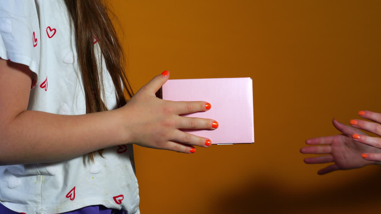 A child hands over a small, pastel pink gift box to another person against a warm orange background, with both sets of hands visible and nails painted in matching orange. cheerful, celebratory Moment.