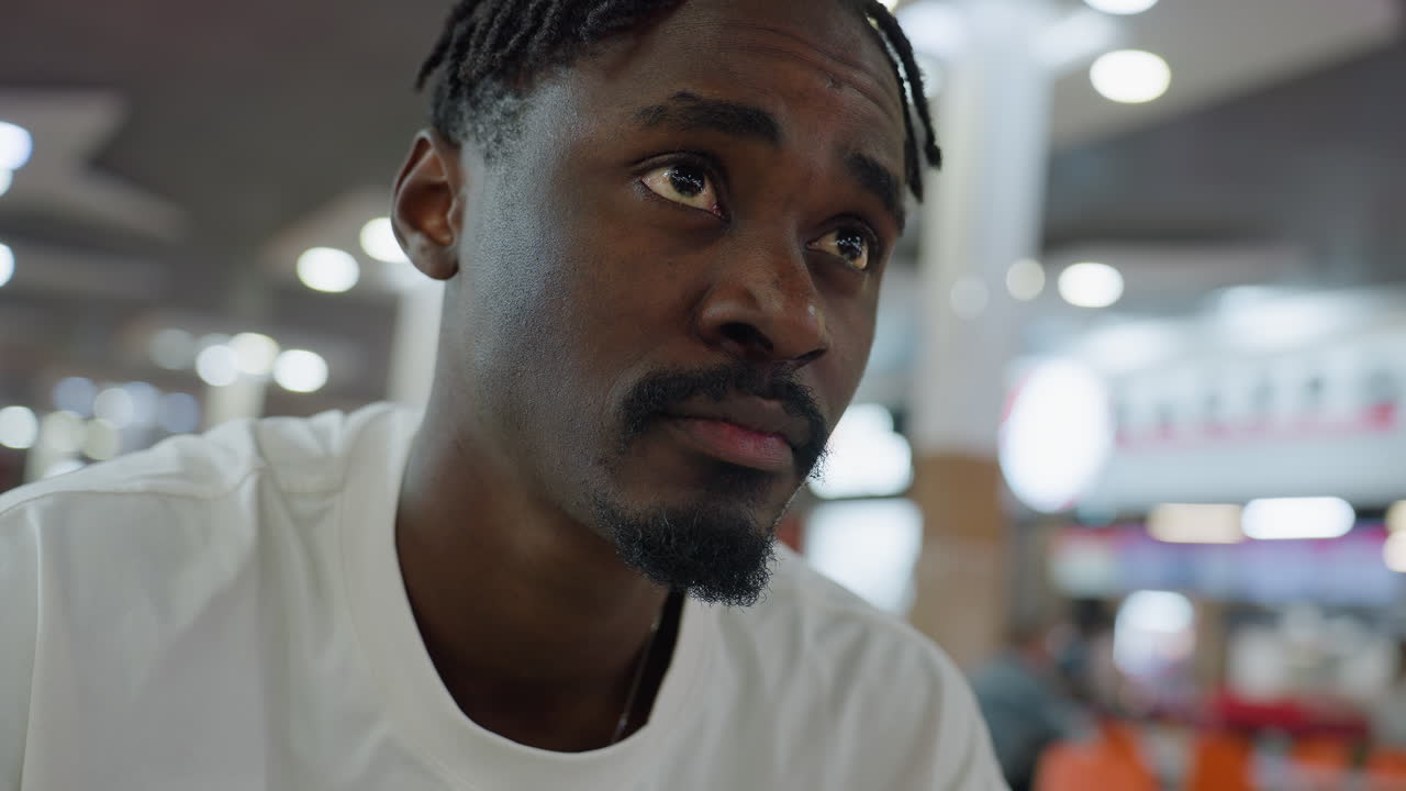 Young african man with braided hair and goatee wearing white shirt sits indoors looking around with contemplative expression, surrounded by blurred bright lights and background
