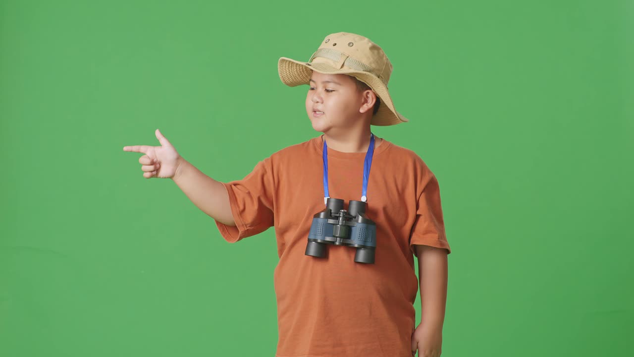 niño turista asiático con un sombrero y binoculares sonriendo y señalando hacia un lado mientras está de pie en el fondo de la pantalla verde. niño investigador examina algo, concepto de aventura de turismo de viaje