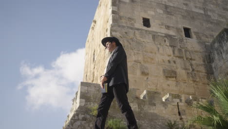 a man in traditional attire stands thoughtfully in front of an ancient fortress in jerusalem, with the towering stone walls creating a powerful backdrop against the clear sky