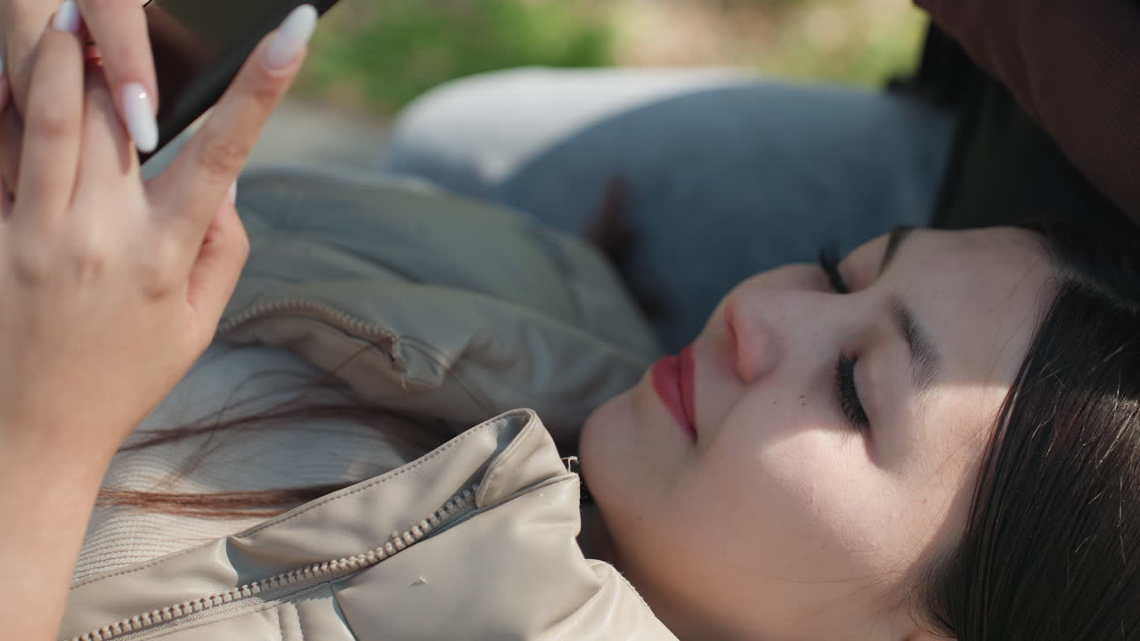 편안한 표정으로 기대앉아 휴대폰 피드를 탐색하는 아시아 여성, 디지털 스크롤과 편안한 자연이 어우러진 느긋한 야외 풍경, 캐주얼 재킷, 부드러운 빛, 사색적인 분위기