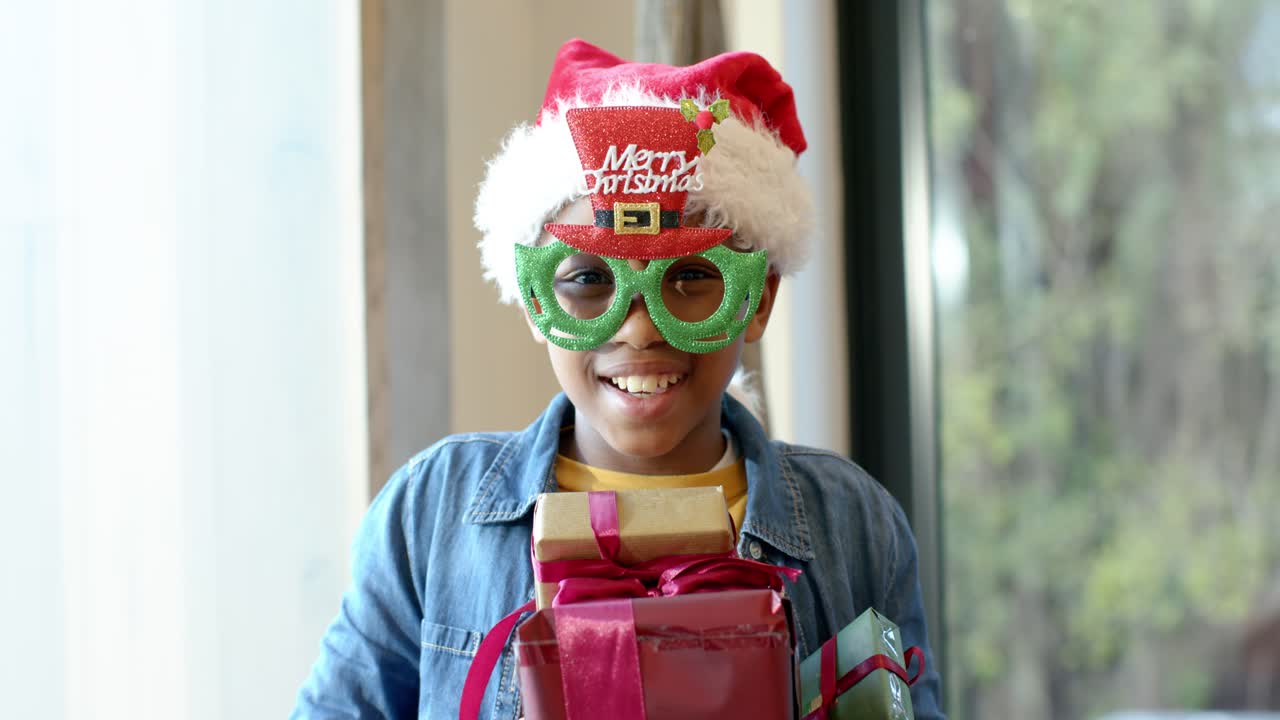 Recording starting, boy holding gifts donning glasses, Santa hat near window, smiling for Christmas
