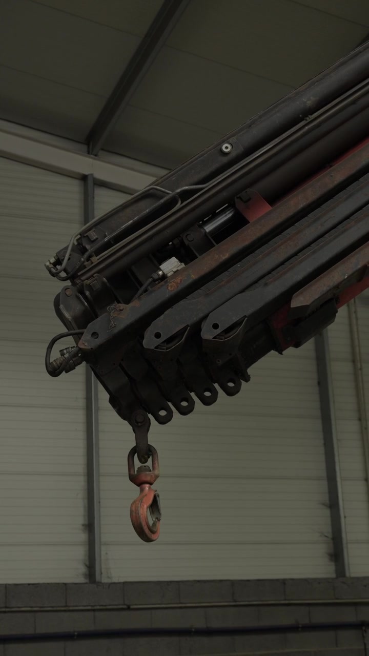 Folding hydraulic knuckle boom crane arm with a suspended hook inside an industrial workshop