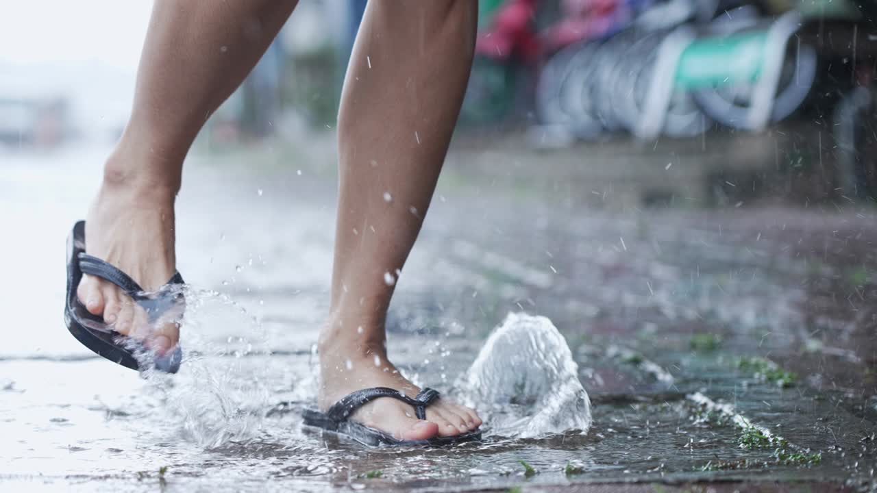 los pies disparados en ángulo bajo en zapatillas pisando pisos de agua en un día de lluvia causado por el monzón, disfruta saltando y salpicando gotas de agua alrededor de la pasarela pavimentada, cámara lenta, cambios climáticos tropicales