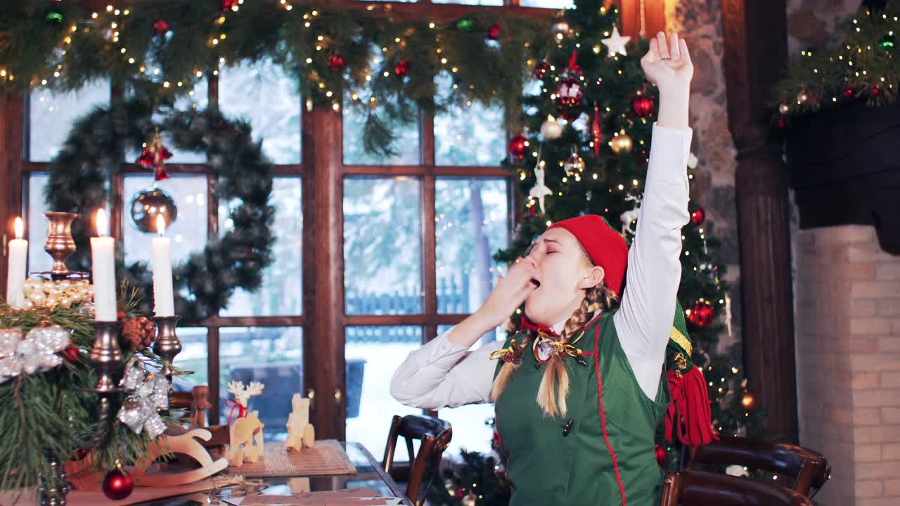A little girl in a green suit wakes up and yawns. Santa claus residence with christmas tree and decorations