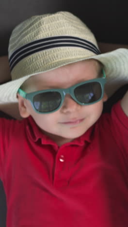 un niño pequeño con elegantes gafas de sol y sombrero de paja yace en el sofá sonriendo. el niño se relaja en el hotel durante las vacaciones de verano vista cercana superior