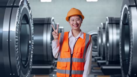 ingeniera asiática con casco de seguridad sonriendo y mostrando un gesto de paz mientras está de pie en una fábrica de metal