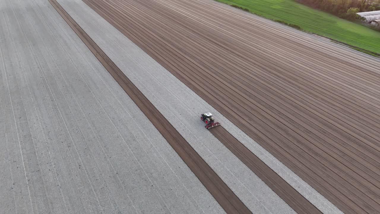 slow orbit around a tractor pulling a harrow as it carves dark seed lines through bleached soil, revealing an endless grid of fresh furrows across a vast agricultural plain under soft daylight