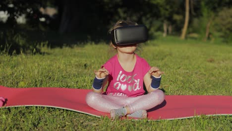 child in vr headset helmet sitting in lotus position on mat and performing yoga meditation in park