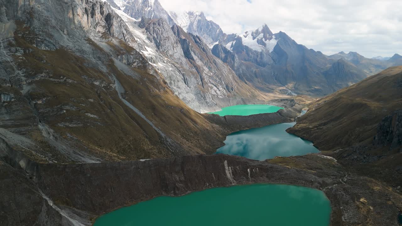 A breathtaking aerial shot tilts up from the three turquoise glacial lakes (Tres Lagunas) to reveal the majestic, snow-capped mountain peaks of the Huayhuash trek in Peru