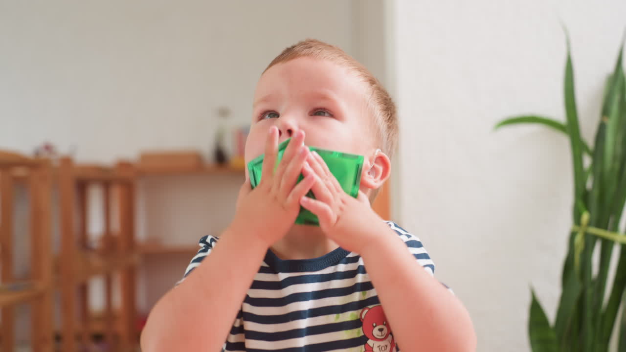 Autistic child in striped shirt holding green toy block close to face while concentrating deeply, wearing hearing aid, seated in bright classroom with blurred wooden furniture background