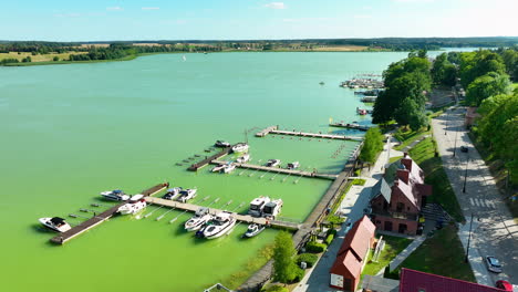 Aerial-view-of-a-marina-with-boats-docked-in-the-green-waters-of-a-lake-in-Ryn,-Warmia-Masuria,-highlighting-the-scenic-lakeside-area