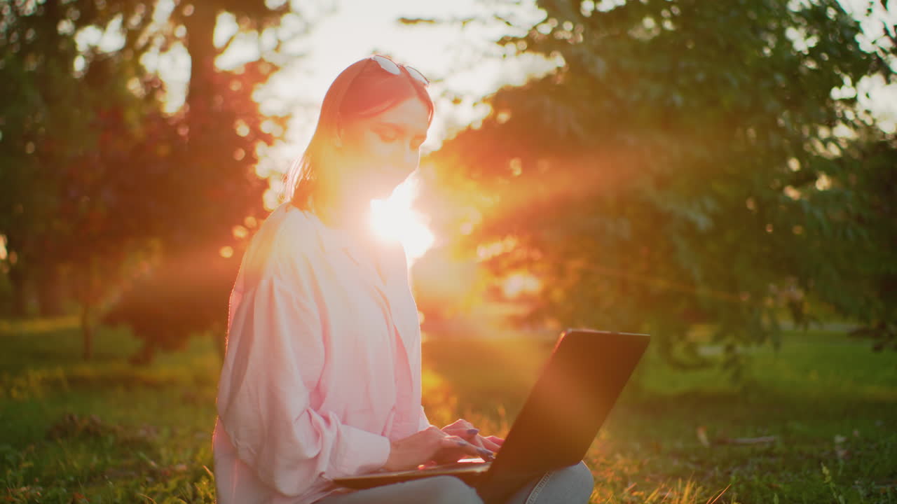 Lady in pink top with glasses resting on her head smiles warmly while looking up thoughtfully and then focusing back on her laptop, beautiful sunlight creates a glowing aura around her outdoors