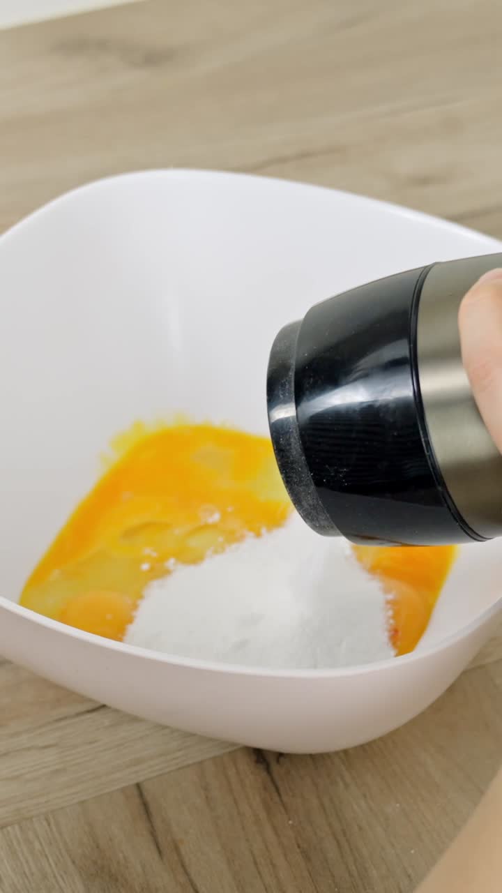 Vertical frame captures powdered sugar being added to golden egg yolks in a white bowl on a wooden surface, a key step in preparing creamy dessert bases like tiramisu or custards
