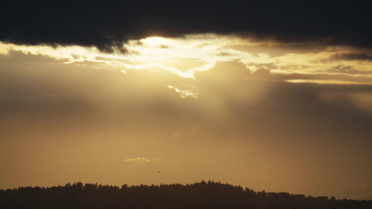 Strong sun beams break through a dark evening cloud band, casting hazy light over a distant forest ridge in Norway.