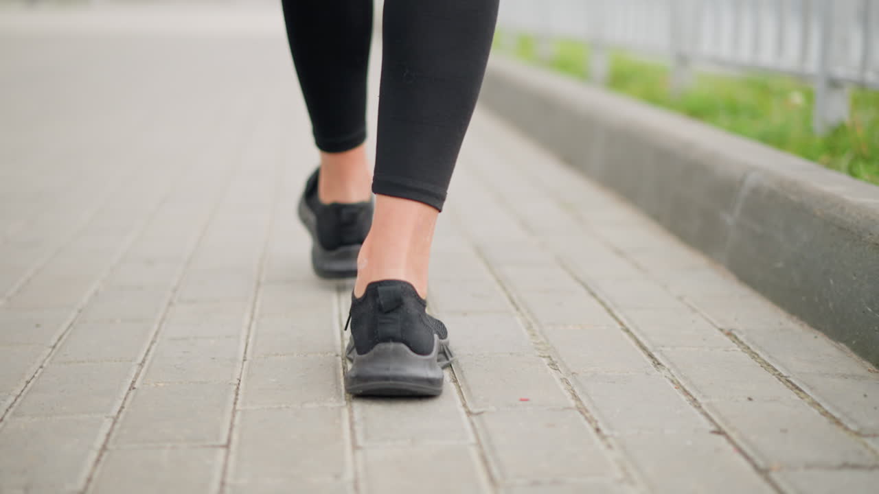 Back view of lady walking majestically in black canvas shoes with focus on her legs and steps, strolling on a smooth pavement beside iron fence and green grass path