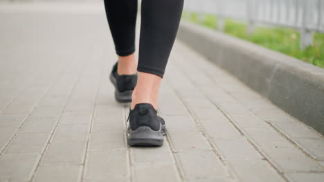 back view of lady walking majestically in black canvas shoes with focus on her legs and steps, strolling on a smooth pavement beside iron fence and green grass path