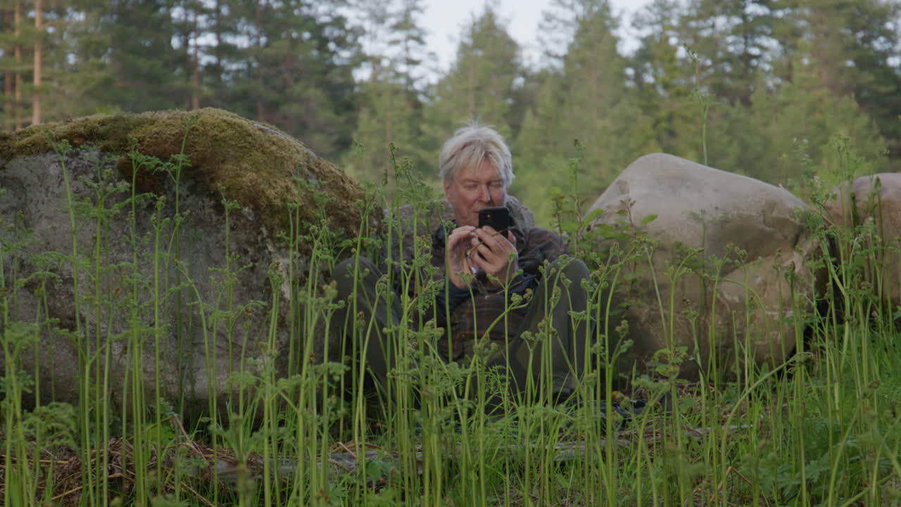Elderly man sitting on forest floor between moss-covered boulders using smartphone, surrounded by tall young fern shoots, expressing themes of solitude, connection and modern nature immersion.
