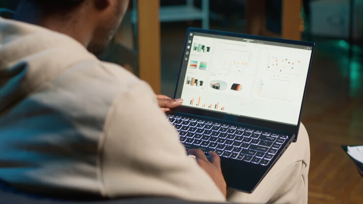 Remote worker accountant doing math calculations using financial graphs on laptop, working at home. Self employed guy in loft comparing business economic figures on notebook. Camera A.