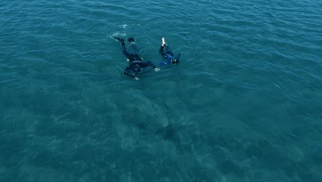 two people wearing wetsuits on crystal blue ocean water surface swim together at villarino beach