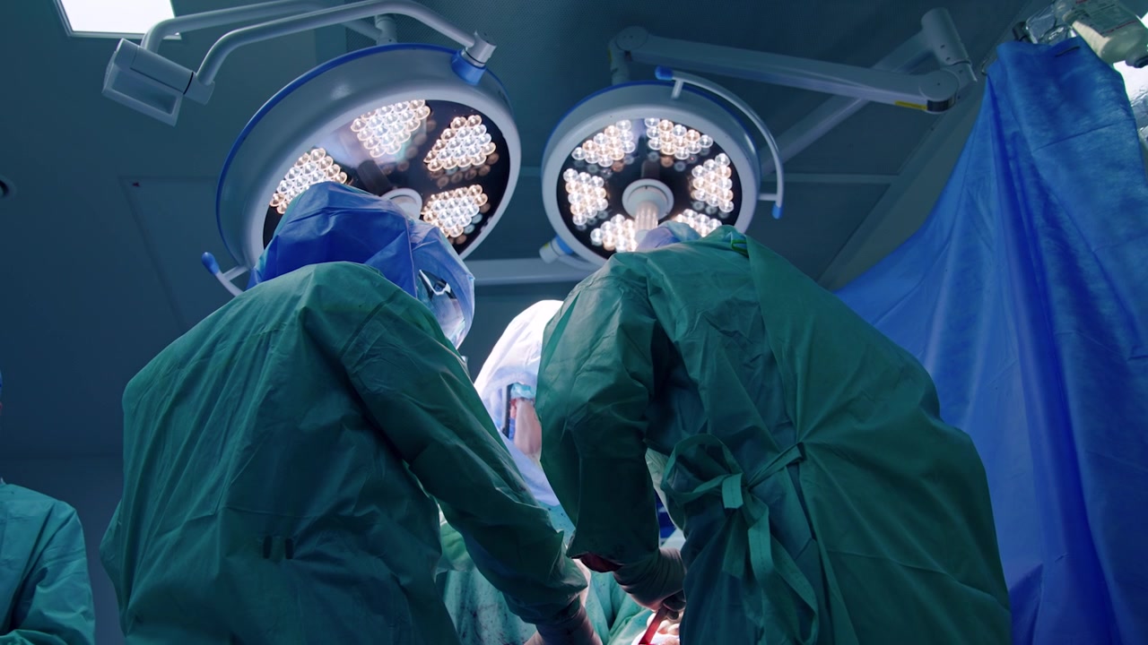 Tre chirurghi sono al tavolo operatorio. Medici con caschi eseguono un intervento chirurgico per traumi sotto le lampade operatorie rotonde. Vista dal basso.