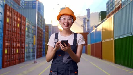 trabajadora asiática con gafas de protección y casco de seguridad sonriendo y señalando el teléfono inteligente en su mano mientras está de pie en el almacén del patio de contenedores
