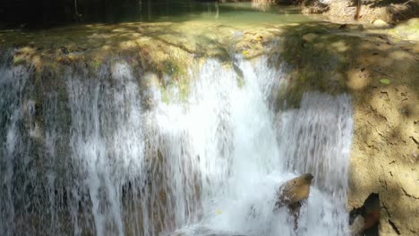 Fresh-clear-white-water-cascades-down-small-rock-wall-in-natural-forest-by-big-stone-in-river,-close-up-pan-up