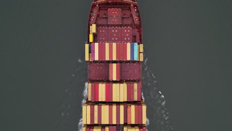 a top down view directly above a large, red container ship with red and yellow containers on the hudson river on a cloudy day