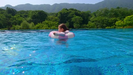 a little girl spins and plays in a pink inflatable tube in a blue pool, surrounded by lush green forest and distant mountains on a sunny day