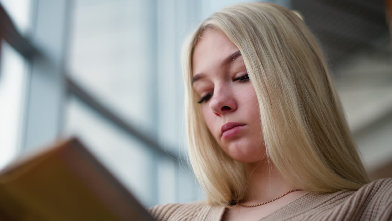 mujer joven enfocada con cabello rubio leyendo su libro en un entorno relajado, perdida en sus pensamientos con un ligero borrón en el fondo, mostrando una sensación de calma y concentración