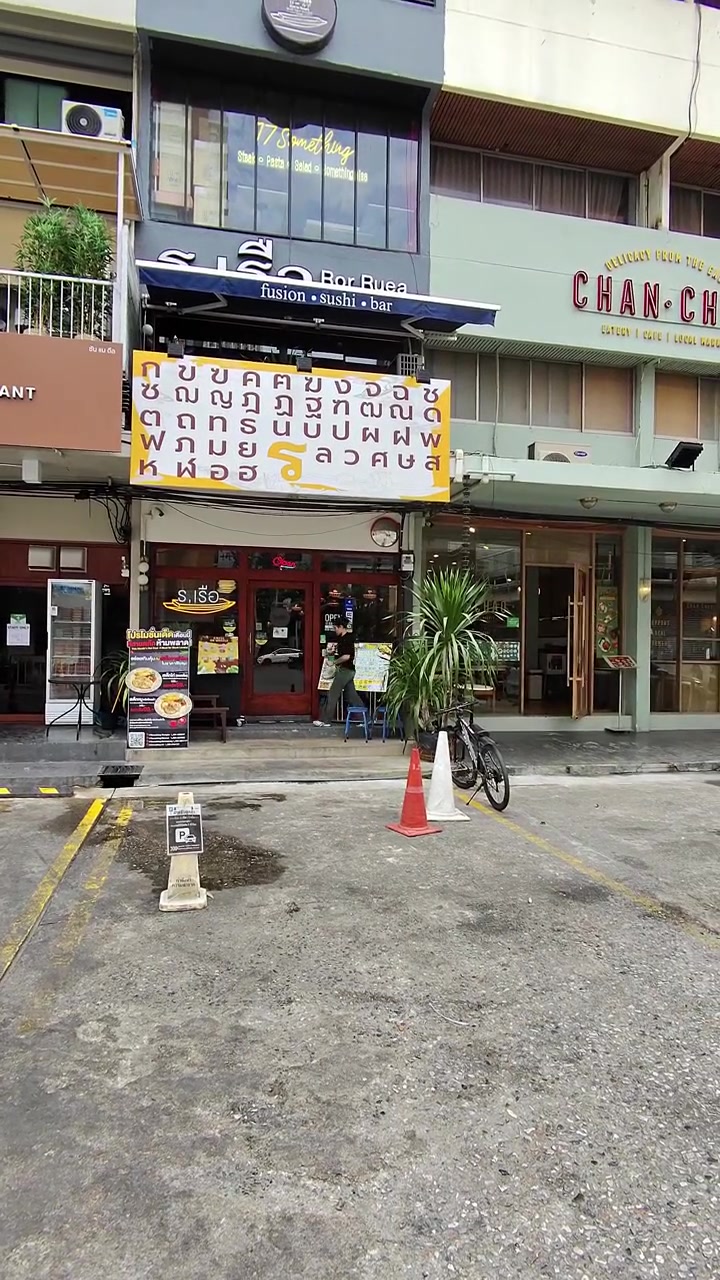 Scatto statico diurno dell'esterno di un ristorante di noodle a Bangkok con coni stradali, striscione del menu e vetrine