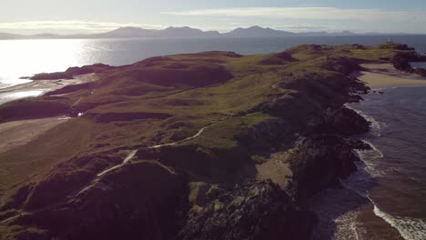 Vista-Aérea-Que-Rodea-La-Isla-Galesa-De-Ynys-Llanddwyn-Con-Un-Océano-Brillante-Y-Una-Brumosa-Cordillera-De-Snowdonia-A-Través-Del-Horizonte-Del-Amanecer.