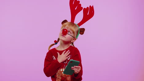 niña pequeña con sombrero de navidad usando teléfono móvil escribiendo una nueva publicación en la web, comprando en línea, navegando