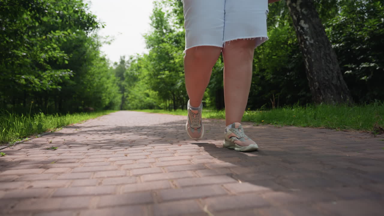 Vista en ángulo bajo de una chica regordeta caminando por un sendero entrelazado en un jardín verde, la luz del sol y las sombras de los árboles se proyectan maravillosamente en el suelo, un edificio distante se ve suavemente a través del follaje