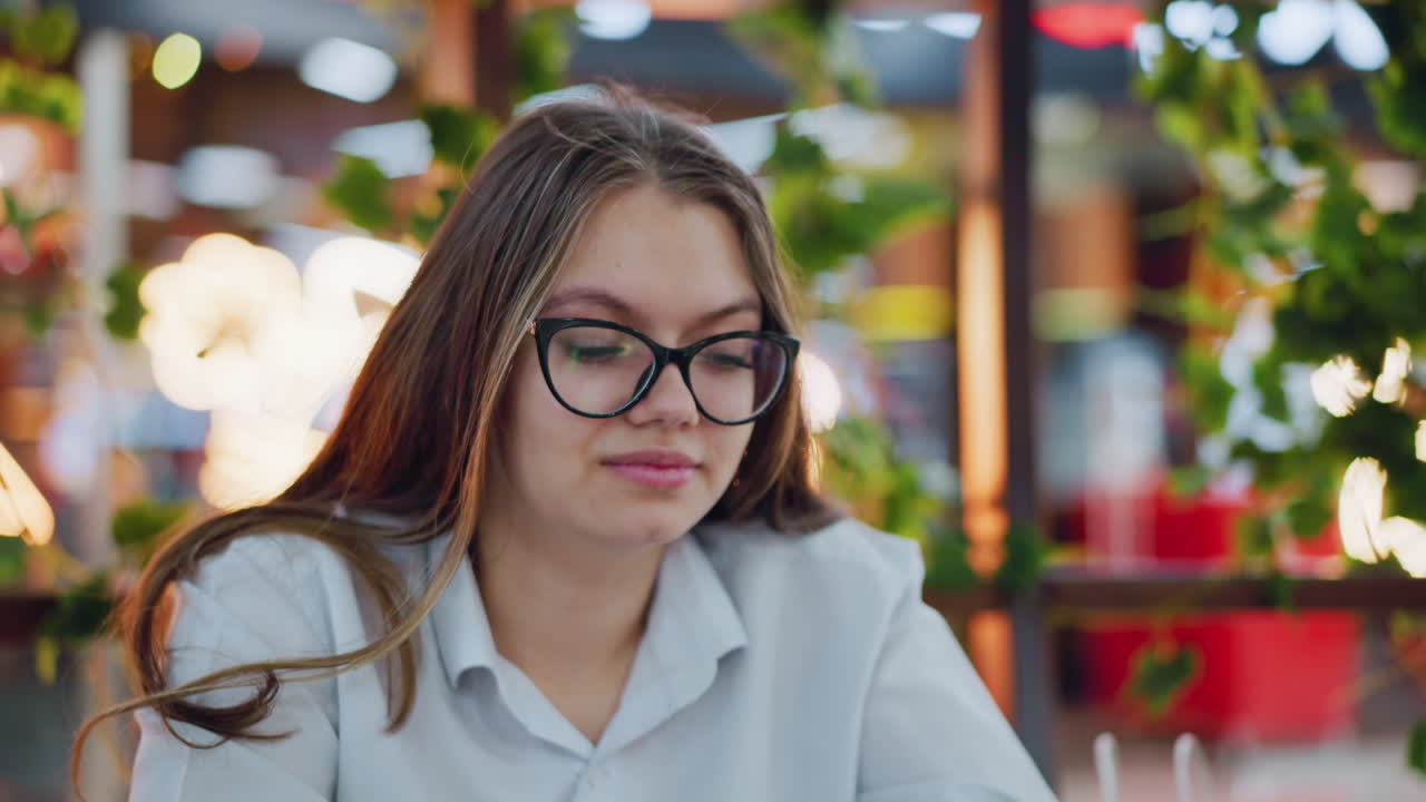 primer plano de una mujer elegante sonriente con gafas, enfocada en su teléfono mientras lo opera, el fondo está decorado con plantas y luces brillantes en un entorno interior elegante