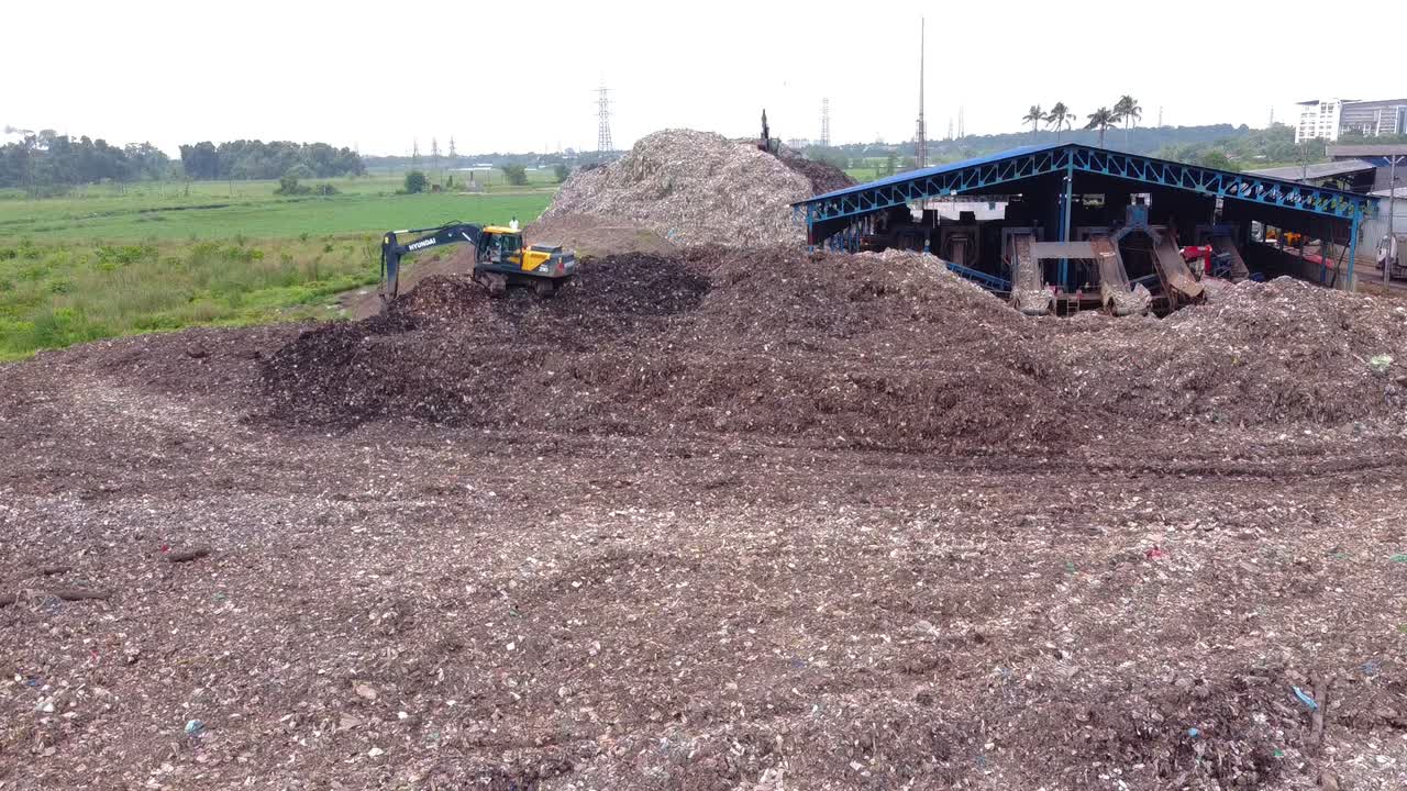 Aerial view of a massive garbage dump and recycling facility with an excavator working on a huge mound of mixed waste next to fields and an urban area
