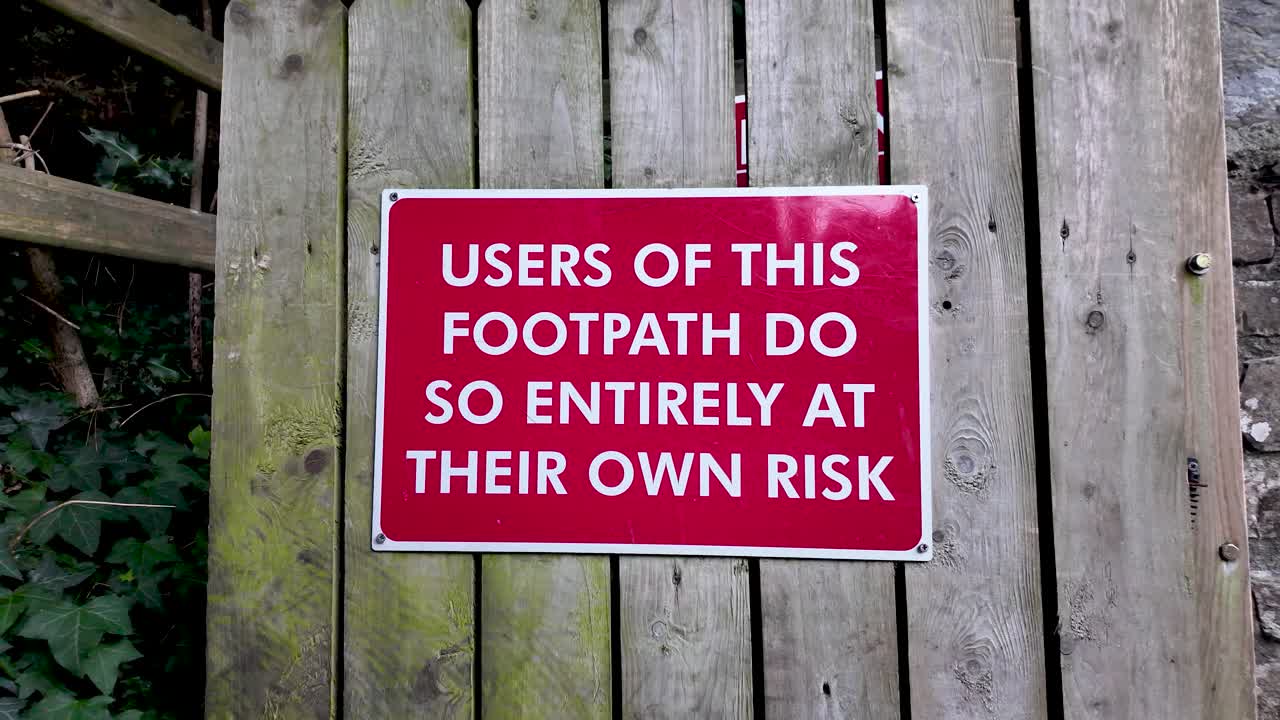 Red warning sign on weathered wood fence, alerting walkers to personal risk on the footpath