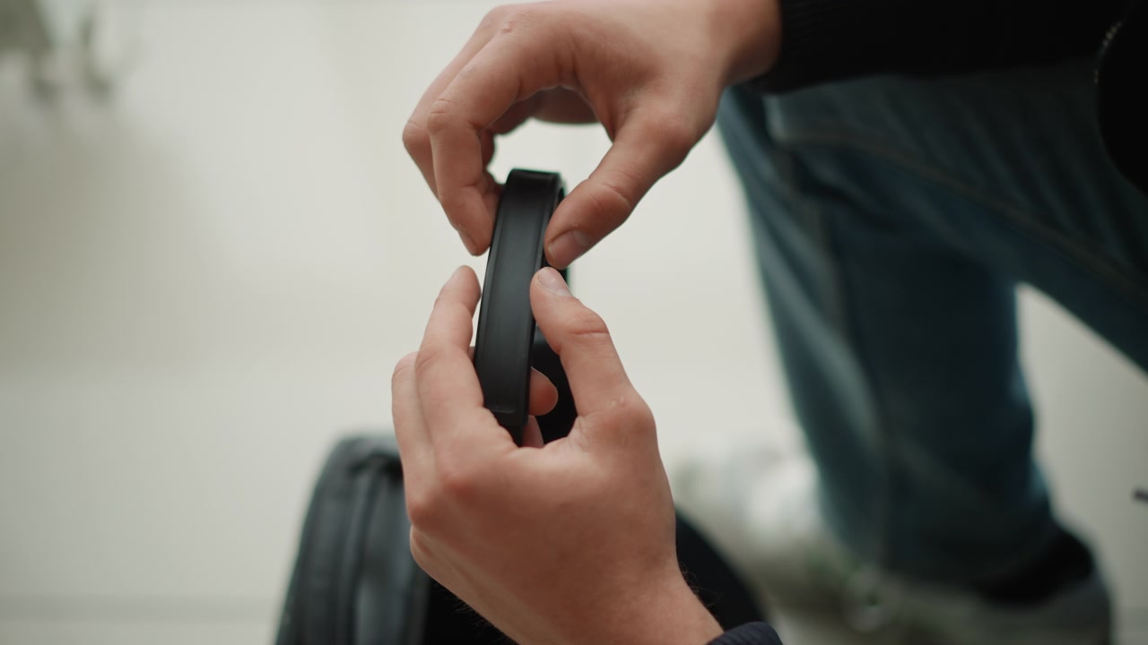 ストラップとイヤーカップに焦点を当て、黒いヘッドホンを調整している手元のクローズアップ。人物はリブ編みの袖口のような質感のディテールが見える暗い色のセーターを着ており、背景は柔らかくぼやけた屋内空間である。