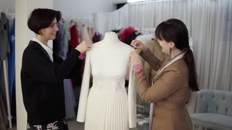 Female-professional-tailors,-designers-working-with-new-model-tailoring-white-dress-on-mannequin-in-studio,-atelier.-Fixing-sleeves-and-pinning-them-from-the-sides.-Fashion-and-tailoring-concept.-Slow-motion