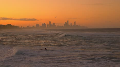 Olas-Del-Océano-Rodando-En-La-Playa-Con-Surfistas---Puesta-De-Sol-Naranja-Con-El-Horizonte-Del-Paraíso-De-Los-Surfistas---Costa-Dorada,-Queensland,-Australia