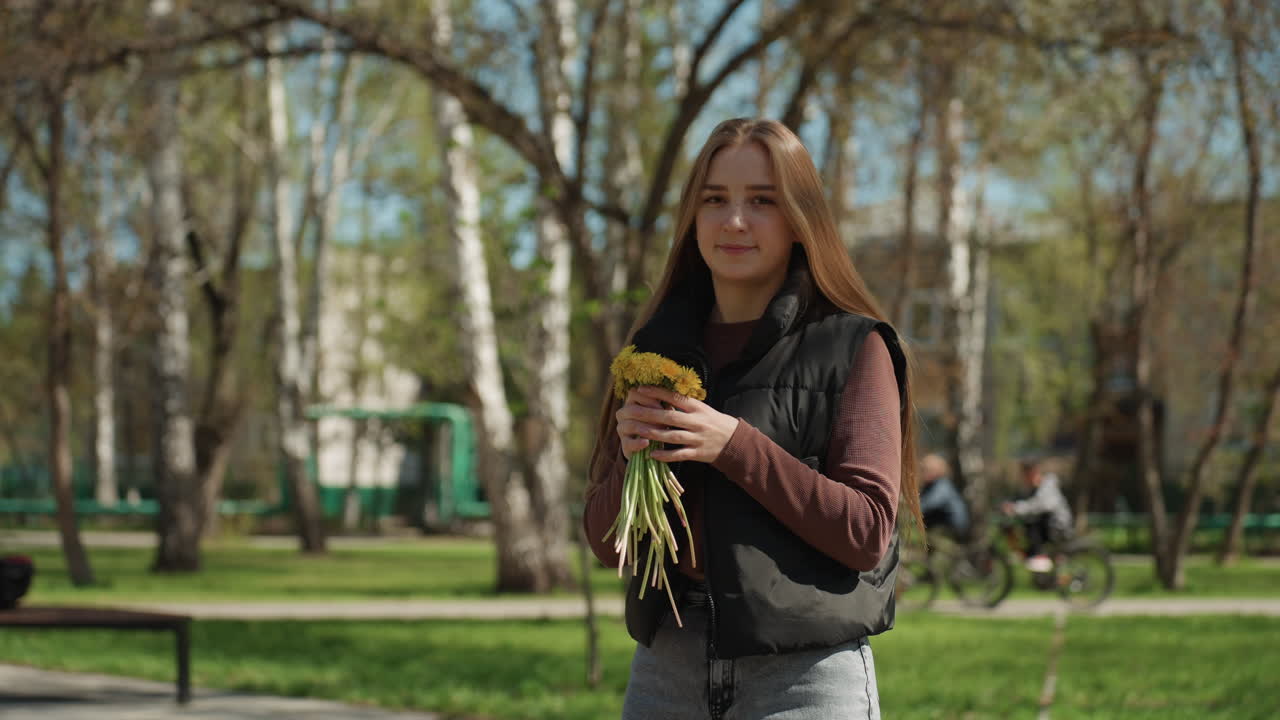 젊은 백인 여성이 자작나무 숲에서 야생화를 향기 맡고, 눈을 감고 차분한 표정으로 작은 노란색 꽃다발을 얼굴 가까이 든 모습; 은은한 향기, 따뜻한 봄, 캐주얼한 조끼와 청바지